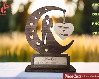 May include: A wooden wedding decoration featuring a silhouette of a couple embracing within a crescent moon shape. A heart-shaped tag with the names "William" and "Emma" hangs from the moon. The base has the text "Nice Cuts - Original Laser Ready Design".