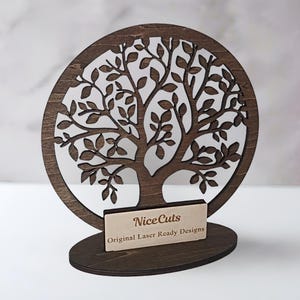May include: A wooden tree of life cutout with a circular design. The tree has a detailed design with many branches and leaves. The tree is mounted on a wooden base with a small sign that reads "NiceCuts Original Laser Ready Designs".