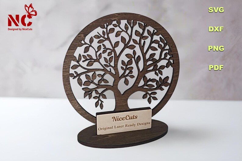 May include: A wooden tree of life cutout with a circular design. The tree has a detailed design with many branches and leaves. The tree is mounted on a wooden base with a small sign that reads "NiceCuts Original Laser Ready Designs".