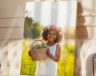 May include: A framed canvas print showing a young girl with a floral crown and a basket of flowers. The girl is smiling, wearing a white dress, and standing in a field of green grass. The print is displayed on a cream-coloured sofa.