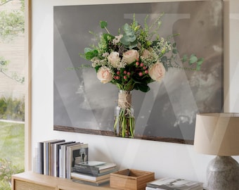 May include: A bouquet of light pink roses, greenery, and berries in a clear glass vase tied with twine. The floral arrangement is displayed on a wooden shelf against a grey backdrop. The image is part of a home decor scene.