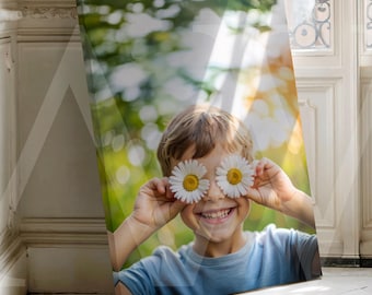 May include: A canvas print showing a smiling child with daisies over their eyes. The child wears a blue top, set against a blurred green and yellow backdrop. The print leans against a white wall, with a window visible.