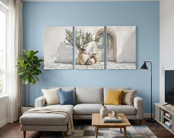 May include: A three-panel canvas wall art set depicting a Mediterranean scene with terracotta pots and an olive tree against a white wall. The artwork is displayed above a light gray sectional sofa with blue and yellow throw pillows in a living room.