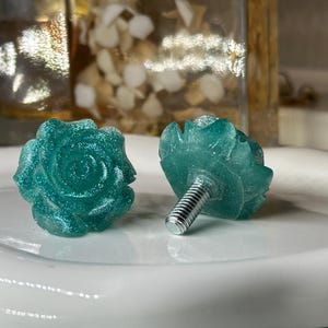 May include: Two teal rose-shaped cabinet knobs with silver screw posts. The knobs have a glittery finish and are set on a white surface. The background includes a blurred display case and an Umbra box.