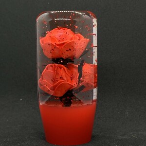 May include: A red gear stick knob featuring three red roses set in clear resin. The base of the knob is solid red, and the resin has red glitter. The knob is cylindrical with a rounded top.