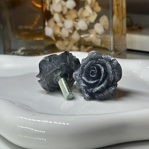 May include: Two dark gray rose-shaped cabinet knobs with silver threaded posts. The knobs are on a white, curved, ceramic tray. The roses have detailed petals and are likely made of stone or a similar material.