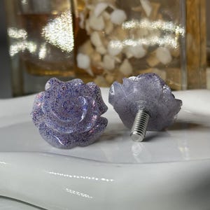 May include: Two lavender rose-shaped cabinet knobs with silver screw posts. The knobs are covered in glitter, with a detailed rose petal design. The knobs are on a white surface, with a blurred background.