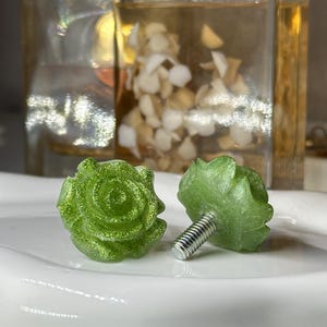 May include: Two green rose-shaped cabinet knobs with glitter accents. One knob is upright, and the other is lying on its side, revealing a silver screw. The knobs are on a white, curved surface.