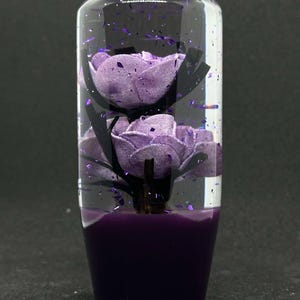 Könnte beinhalten: Ein transparenter Glasschaltknauf mit einem violetten Sockel und zwei violetten Blumen im Inneren. Die Blumen sind von violettem Glitzer umgeben.