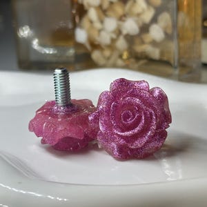 May include: Two decorative cabinet knobs with a rose design. The knobs are a sparkly pink color and have a silver screw base. The knobs are sitting on a white surface.