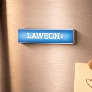 Könnte beinhalten: Ein rechteckiger Magnet mit dem Wort "LAWSON" in weißen Großbuchstaben auf blauem Hintergrund. Die japanischen Schriftzeichen befinden sich in der unteren rechten Ecke. Der Magnet haftet auf einer metallischen Oberfläche.