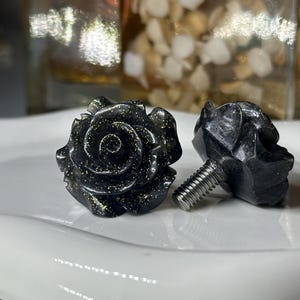 May include: Two black rose-shaped cabinet knobs with gold flecks. One knob is fully assembled, showing the threaded screw. The other knob is partially visible, displaying the rose design and screw.
