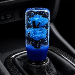 May include: A cylindrical gear shift knob made of clear resin containing two blue roses and blue glitter flakes. The base of the knob is a solid blue colour, and it is mounted onto a black leather gear shift boot inside a vehicle interior.