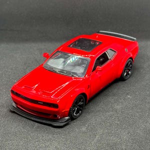 May include: A bright red die-cast model car featuring a black front splitter, black wheels, a sunroof, and a rear spoiler. The vehicle has a sleek, two-door coupe body style with a detailed front grille and side mirrors, captured from a high angle.