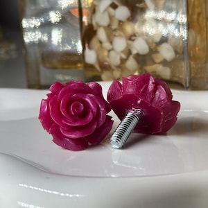 May include: Two magenta rose-shaped cabinet knobs with silver screw threads. The knobs are on a white, slightly curved surface. The background includes a blurred glass container and other decorative items.