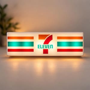 May include: A rectangular light box featuring the 7-Eleven logo. The logo includes the word "ELEVEN" in teal, with the number 7 in red and orange. The light box has horizontal stripes in orange, teal, and red.