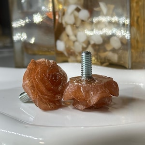 May include: Two rose-shaped cabinet knobs. The knobs are a translucent orange with gold flecks and a silver screw. The knobs are on a white, scalloped plate. The background is blurred.