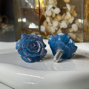 May include: Two blue rose-shaped cabinet knobs with red glitter accents. The knobs have a screw base for easy installation. The knobs are on a white surface. Decorative hardware for furniture.