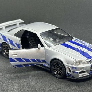 Op de afbeelding: Een zilveren speelgoedauto met blauwe en witte race strepen. De auto heeft een zwart interieur en een spoiler op de achterkant. De auto is een Nissan Skyline GT-R R34.