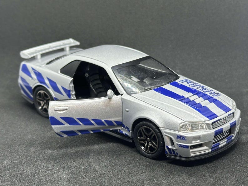 Op de afbeelding: Een zilveren speelgoedauto met blauwe en witte race strepen. De auto heeft een zwart interieur en een spoiler op de achterkant. De auto is een Nissan Skyline GT-R R34.