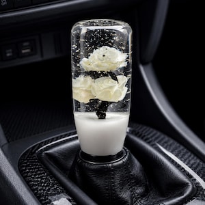 May include: A cylindrical gear shift knob made of clear resin containing two white flowers with dark stems and gold flecks. The base of the knob is opaque white, and it is mounted on a black leather gear shift boot inside a vehicle interior.