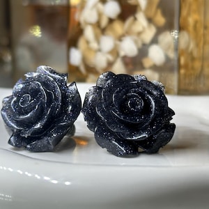May include: Two dark blue rose-shaped sculptures with sparkling inclusions sit on a white surface. The detailed carvings of the rose petals are visible. The background is blurred, suggesting a focus on the floral sculptures.
