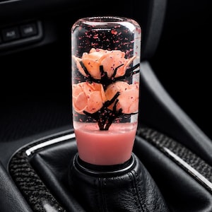 May include: A cylindrical gear shift knob made of clear resin containing three peach-coloured flower blossoms and black branch-like structures. The base of the knob is a solid opaque pink, and the interior is filled with small, scattered red flakes.