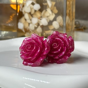 May include: Two magenta rose-shaped earrings with glitter accents sit on a white, wavy-edged plate. The earrings are the focal point, showcasing a detailed floral design. The background includes a glass container and decorative elements.