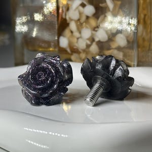 May include: Two dark blue rose-shaped cabinet knobs with a sparkling, glittery appearance. One knob is upright, while the other lies on its side, revealing a silver screw. The knobs are on a white, glossy surface.