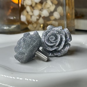 May include: Two silver glitter rose-shaped stud earrings. The earrings are on a white, slightly curved surface. The roses have detailed petals and a metallic sheen. The earring posts are visible, suggesting a jewelry item.