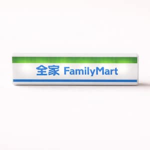 Puede incluir: Un letrero rectangular con el texto "全家 FamilyMart" en azul y blanco. La parte superior del letrero es verde y la inferior tiene una franja azul. El letrero está sobre un fondo blanco.
