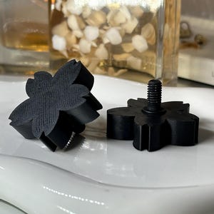 May include: Two black, flower-shaped objects. One is lying on its side, and the other has a threaded post extending from the center. The objects are on a white, oval-shaped plate. The background includes a glass bottle and a white box.