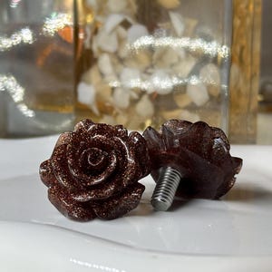 May include: Two rose-shaped cabinet knobs. The knobs are a dark reddish-brown color with glittery accents. Each rose has detailed petals and a silver screw post for mounting. The knobs are on a white surface.