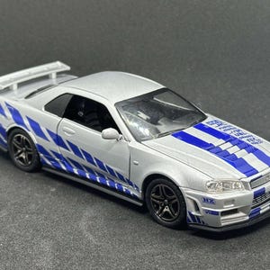 May include: A silver Nissan Skyline GT-R R34 model car with blue and white racing stripes. The car has a black interior and a rear spoiler.
