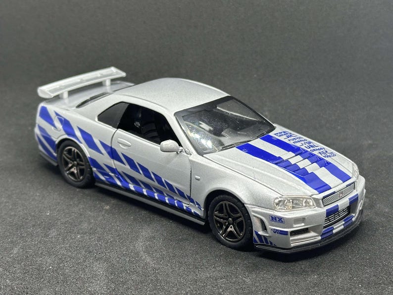 Op de afbeelding: Een zilveren Nissan Skyline GT-R R34 model auto met blauwe en witte race strepen. De auto heeft een zwart interieur en een achter spoiler.