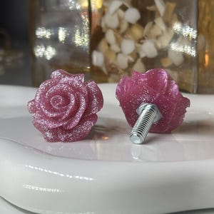 May include: Two rose-shaped cabinet knobs. The knobs are a deep pink color with silver glitter accents. One knob is shown with a silver screw attached. The knobs are sitting on a white, curved surface.