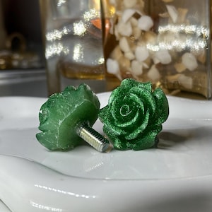May include: Two green rose-shaped cabinet knobs with metallic silver screws. The knobs are made of a translucent, glittery material, and are resting on a white, curved surface. The background includes a blurred view of decorative items.
