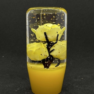 May include: A yellow and clear resin shift knob with a floral design. The shift knob features three yellow flowers with a dark brown stem and gold flakes suspended in the clear resin. The base of the shift knob is solid yellow.