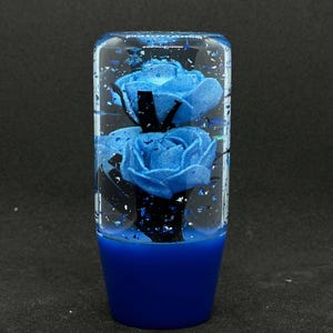 May include: A blue shift knob featuring two blue roses set in clear resin. The base of the knob is solid blue. Silver glitter flakes are suspended in the resin. This is a decorative car accessory.
