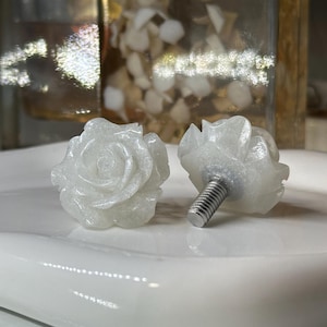 May include: Two white rose-shaped cabinet knobs. The knobs are carved with detailed petals and have a silver screw post. The knobs are displayed on a white, glossy surface.