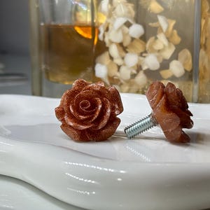 May include: Two rose-shaped cabinet knobs in a reddish-brown hue rest on a white, curved ceramic dish. The knobs have a detailed floral design and a silver screw post. The background includes a glass and dried flowers.
