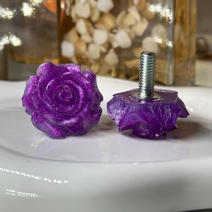 May include: Two purple rose-shaped cabinet knobs with glitter. One knob is fully formed, while the other has a metal screw attached. The knobs are on a white surface, with blurred background elements.