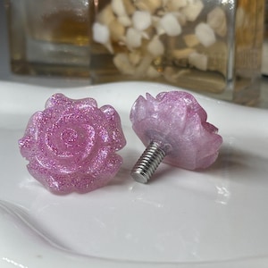 May include: Two decorative rose-shaped cabinet knobs. The knobs are a light pink color with glitter and a silver screw. The knobs are on a white surface. The knobs are a decorative hardware item.