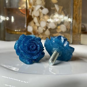 May include: Two blue rose-shaped cabinet knobs with a glittery finish. The knobs are made of a translucent material and have a silver screw for attachment. The knobs are on a white surface.