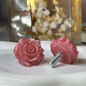May include: Two rose-shaped cabinet knobs in a shimmering red hue. The knobs are made of a translucent material with visible glitter, and each has a silver screw for attachment. They are displayed on a white, scalloped plate.