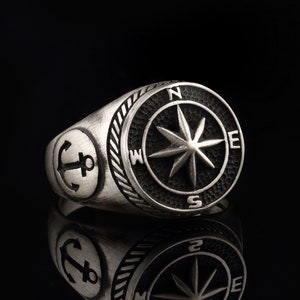 Puede incluir: Un anillo de plata con un diseño de brújula y ancla. La cara de la brújula es negra con una estrella plateada y las letras N, S, E y W. El ancla está en el lado del anillo.