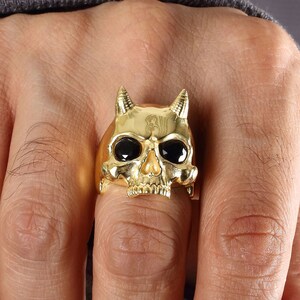 Puede incluir: Un anillo de calavera de oro con ojos de gemas negras y cuernos. El anillo se lleva en un dedo.