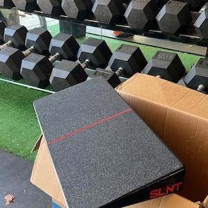 May include: A black and grey exercise slant board with a red stripe and the word "SLNT" in red. The board is on top of a cardboard box, with a rack of black dumbbells in the background.