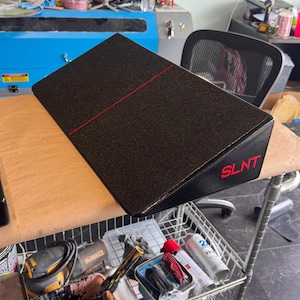 May include: Black, angled, rectangular object with a red line and the word "SLNT" in red. The object is on a brown surface, with tools and supplies visible in the background.