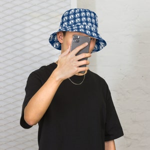 May include: A person wearing a blue bucket hat with a white pattern of circles and a black t-shirt. The person is holding a black smartphone in front of their face.